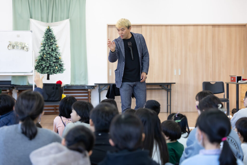 富山のクリスマス会でマジックショー!プロの写真とAIの活用術。 4 児童クラブのステージで、身振り手振りを交えて子供たちに語りかけるマジシャン・コンプレッサー。真剣な眼差しで話に聞き入る子供たちの後姿と、クリスマスの装飾が施された会場の様子。