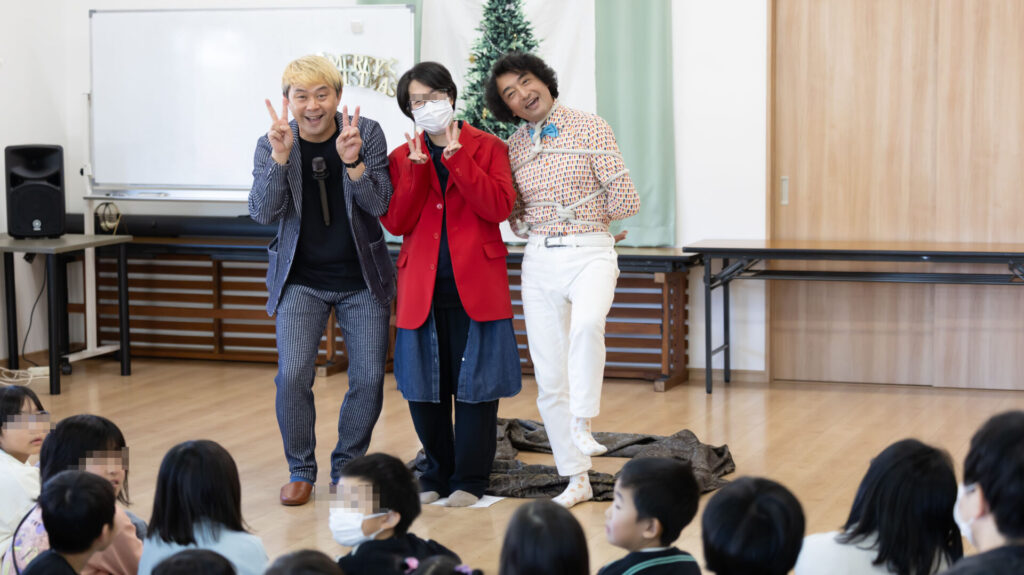 富山のクリスマス会でマジックショー!プロの写真とAIの活用術。 6 クリスマスツリーの前で、プロマジシャンのコンプレッサーと縄で縛られた共演者、そして笑顔の女性が並んでピースサインをしている様子。会場には多くの子供たちが集まり、温かい雰囲気に包まれている。