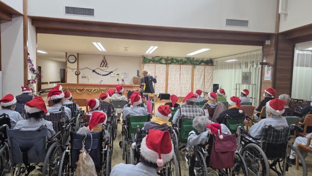 立山町の施設でマジックショー!反応が薄くても「心」は通じ合う! 2 立山町の高齢者施設でのマジックショーの様子。サンタ帽をかぶった利用者様と笑顔で交流するマジシャン・コンプレッサー、そして温かく見守る施設スタッフ。クリスマスの飾り付けがされた会場全体が和やかな雰囲気に包まれている。