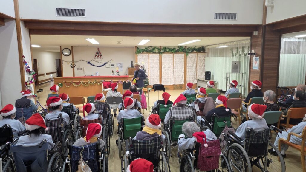 立山町の施設でマジックショー!反応が薄くても「心」は通じ合う! 1 立山町の高齢者施設でのマジックショーの様子。サンタ帽をかぶった利用者様と笑顔で交流するマジシャン・コンプレッサー、そして温かく見守る施設スタッフ。クリスマスの飾り付けがされた会場全体が和やかな雰囲気に包まれている。