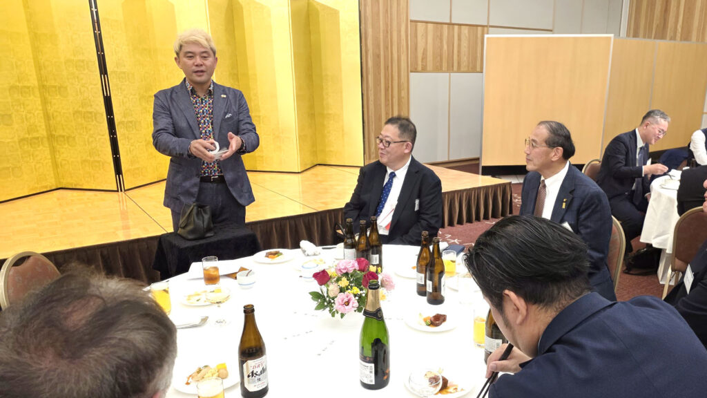 祝賀会のテーブルマジックで、ゲストの目の前でトランプマジックを披露するマジシャン・コンプレッサー。円卓を囲む男性ゲストたちが、至近距離で繰り広げられる不思議な技を興味深く見守っている場面。