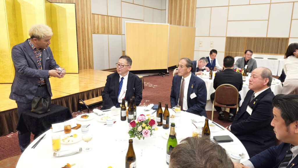 テーブルマジック中に笑顔でゲストに語りかけるコンプレッサー。華やかな柄のシャツに紺のジャケットを合わせ、トランプを手に持ちながら、ホテルアクア黒部の会場内でゲストとの交流を楽しんでいる様子。