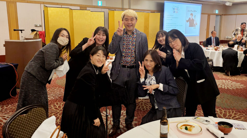 北信ホールディングス様新年会の会場で、ゲストの皆様と笑顔でポーズをとるマジシャンコンプレッサー。温かなご縁を感じる記念写真。