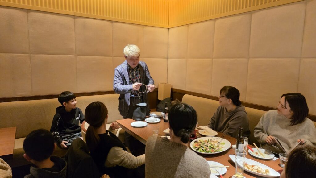 企業新年会でサロンショーを披露するマジシャン・コンプレッサー。立ち上がってリング（金属の輪）を使ったマジックを演じ、お客様が興味深そうに見入っている