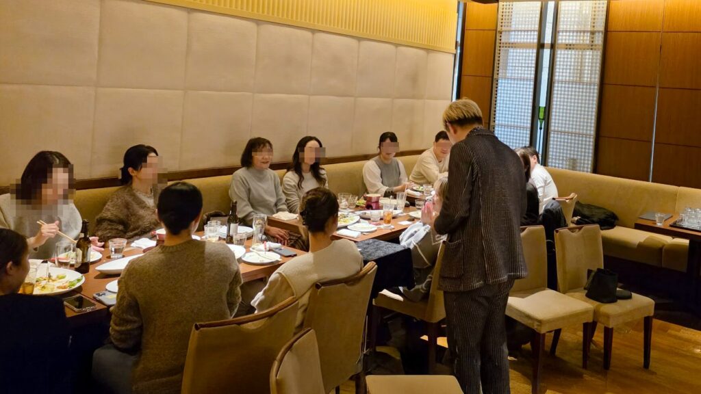 「カジュアルダイニング BON」の新年会会場でパフォーマンス中のマジシャン・コンプレッサー。ストライプスーツ姿で、長テーブルに沿って座るお客様の前でマジックを披露している