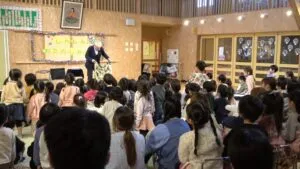 ステージ上でマジシャン・コンプレッサーが和柄のシルク布を顔の前に広げてマジックを演じている瞬間。園児たちが真剣な表情で見つめている。