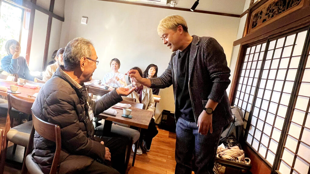 参加者の男性の目の前でマジックを披露するマジシャン・コンプレッサー。至近距離での驚きの瞬間に、周囲の皆様からも笑みがこぼれる、コミュニケーションが生まれている瞬間。