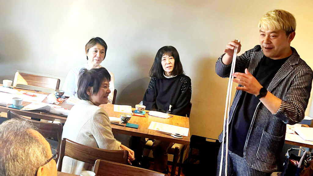 キャリアネットワーク北陸の皆様の前で、白いロープを使ったマジックを実演するマジシャン・コンプレッサー。間近で見る不思議な現象に、参加者の女性たちが興味津々な表情で見入っている様子。