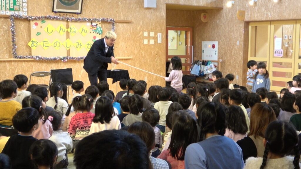 マジシャン・コンプレッサーがロープを使ったマジックを披露し、女の子がお手伝いとしてステージに上がっている場面。たくさんの園児たちが座って見守っている。