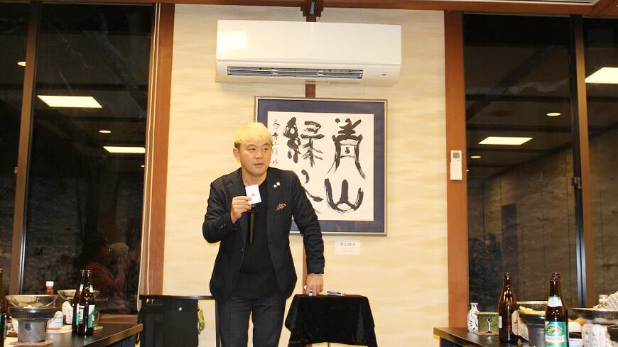 マジシャン・コンプレッサーが宴会場のステージでトランプマジックを披露している様子。金髪に紺のジャケット姿で、手元に1枚のカードを高く掲げている。背景には力強い書が飾られており、和やかな中にも緊張感のあるショーの雰囲気が伝わる。