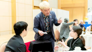 パーティー会場の円卓で、トランプを広げてゲストにカードを選んでもらっているコンプレッサーの様子。