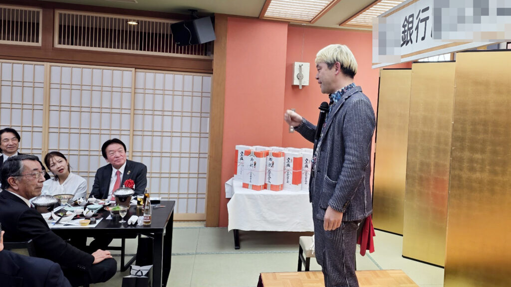 金屏風の前で、派手なスーツを着たマジシャンがマイクを手に笑顔でオープニングトークをしている
金屏風の華やかなステージ。まずは「お金のマジック」からスタートして会場を盛り上げます！