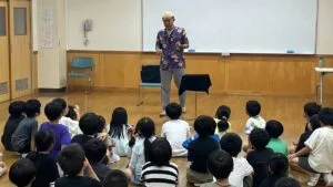 富山県高岡市の学童保育で、床に座った小学生たちの前に立ってマジックショーを進めるマジシャン・コンプレッサー