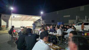 富山県内の企業の秋祭りの夜、屋外テントのステージ前でマジックを見せるマジシャン・コンプレッサーと、長いテーブルで食事をしながら見る皆さん