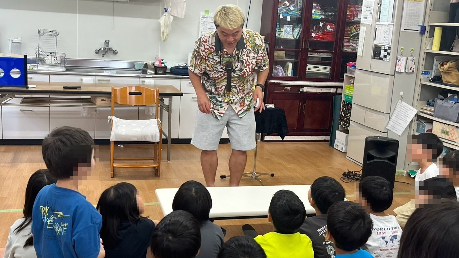 石川県白山市の学童保育で床に座った子どもたちの前に立つマジシャン・コンプレッサー