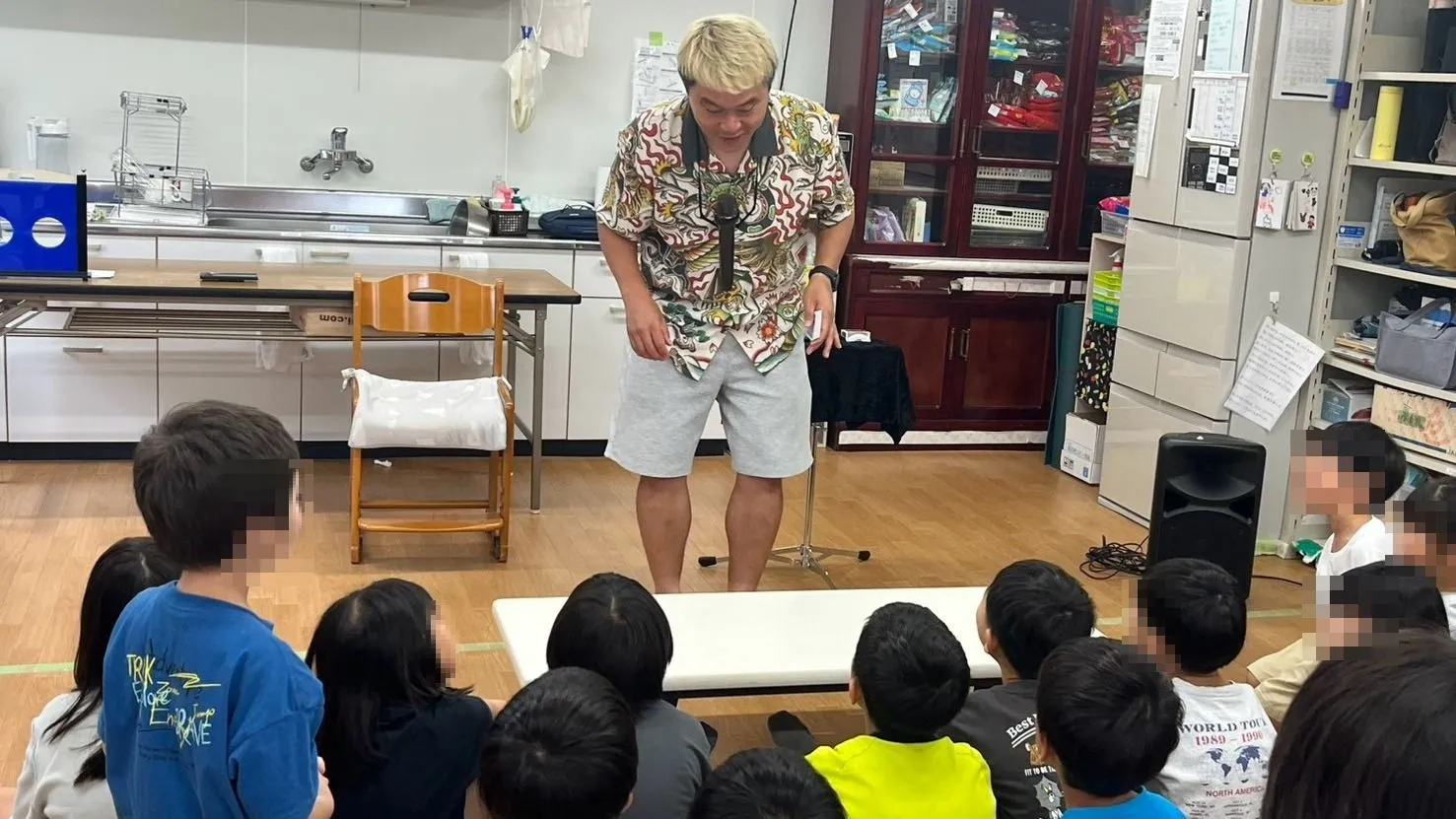 石川県白山市の学童保育で床に座った子どもたちの前に立つマジシャン・コンプレッサー