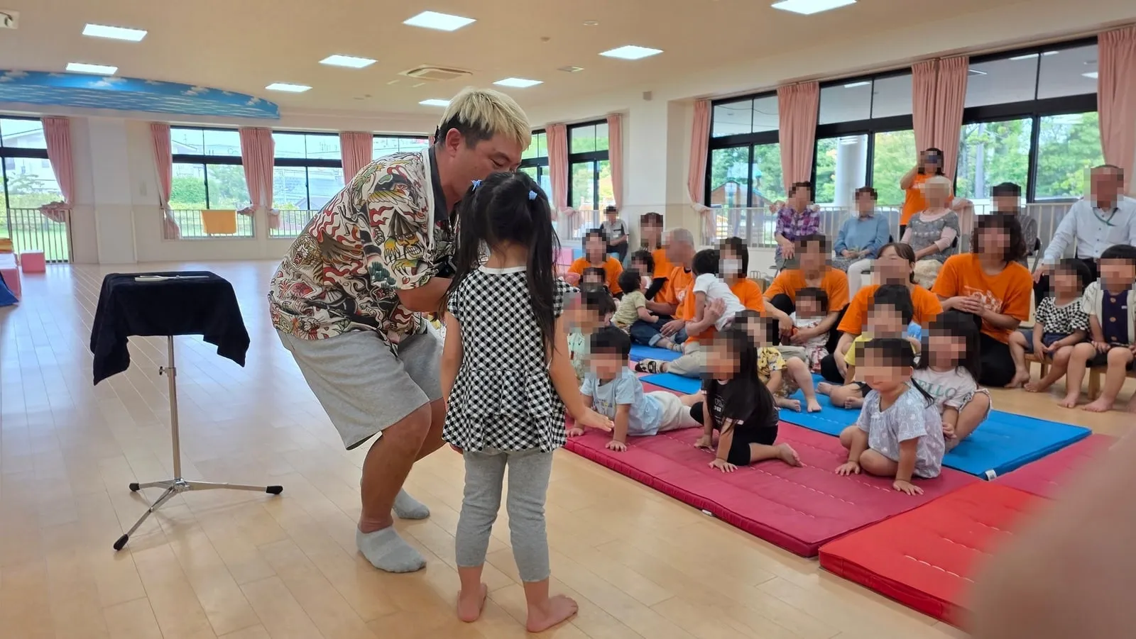 富山市の保育園のプレイルームで女の子と向かい合ってマジックを演じるマジシャン・コンプレッサーと見つめる園児さんたち