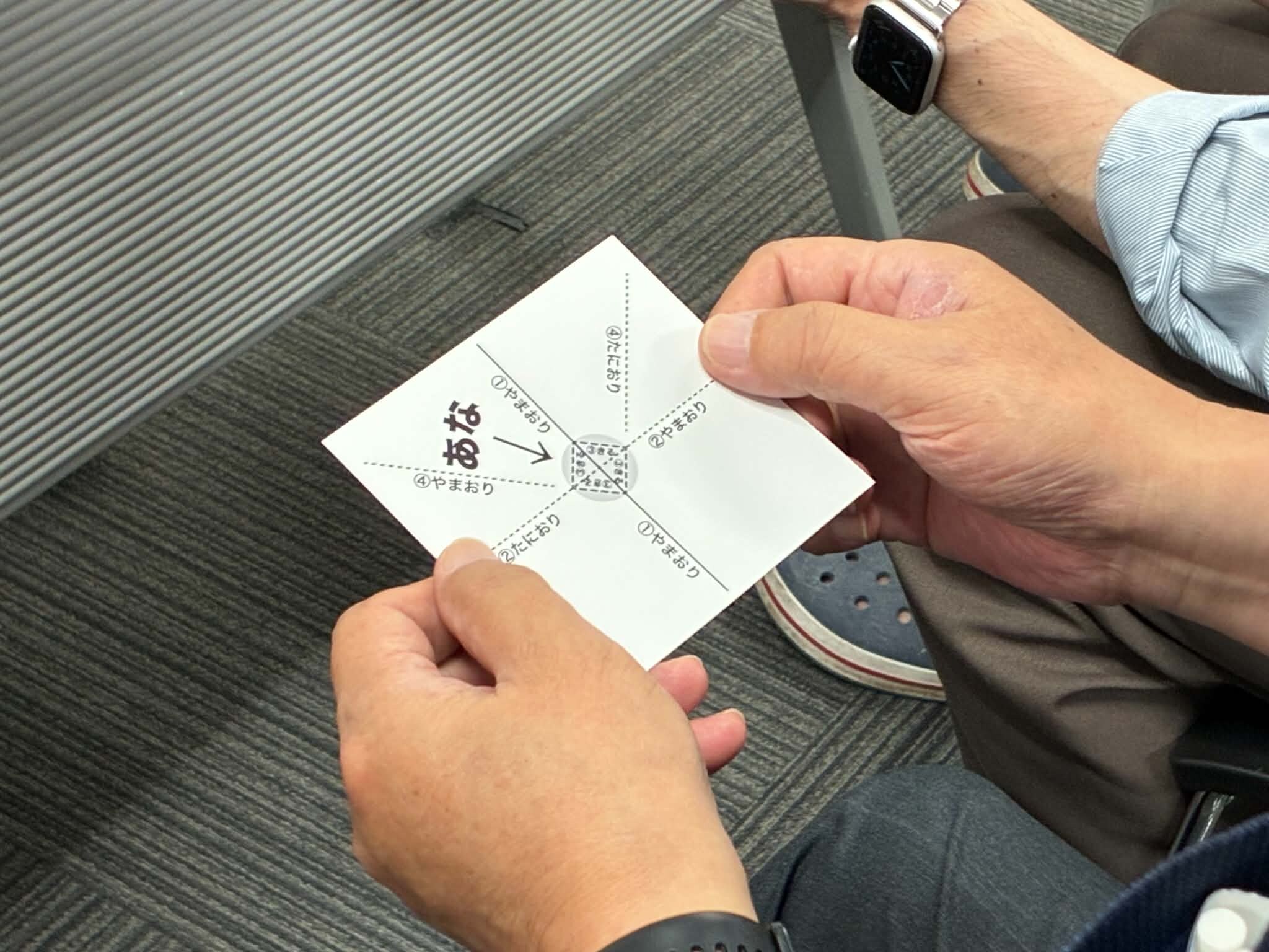 「あな」と書かれた厚手の紙に、山折り・谷折りの線が入った紙のマジックを手に取るまじっぱのメンバー｜射水市のマジックサークル