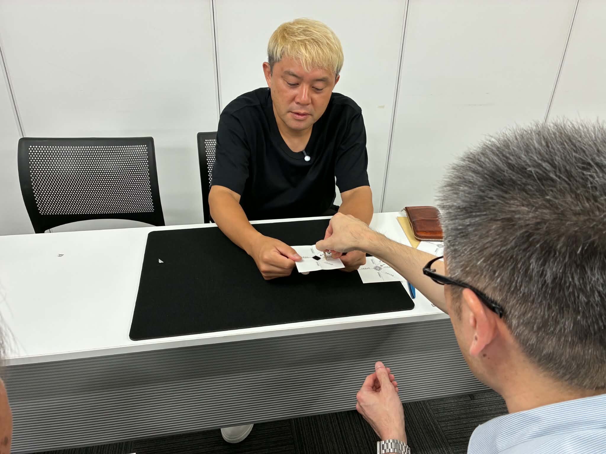 マジシャン・コンプレッサーが紙のマジックを見せ、メンバーが500円玉を穴に通そうと手をのばす様子｜まじっぱ130回目