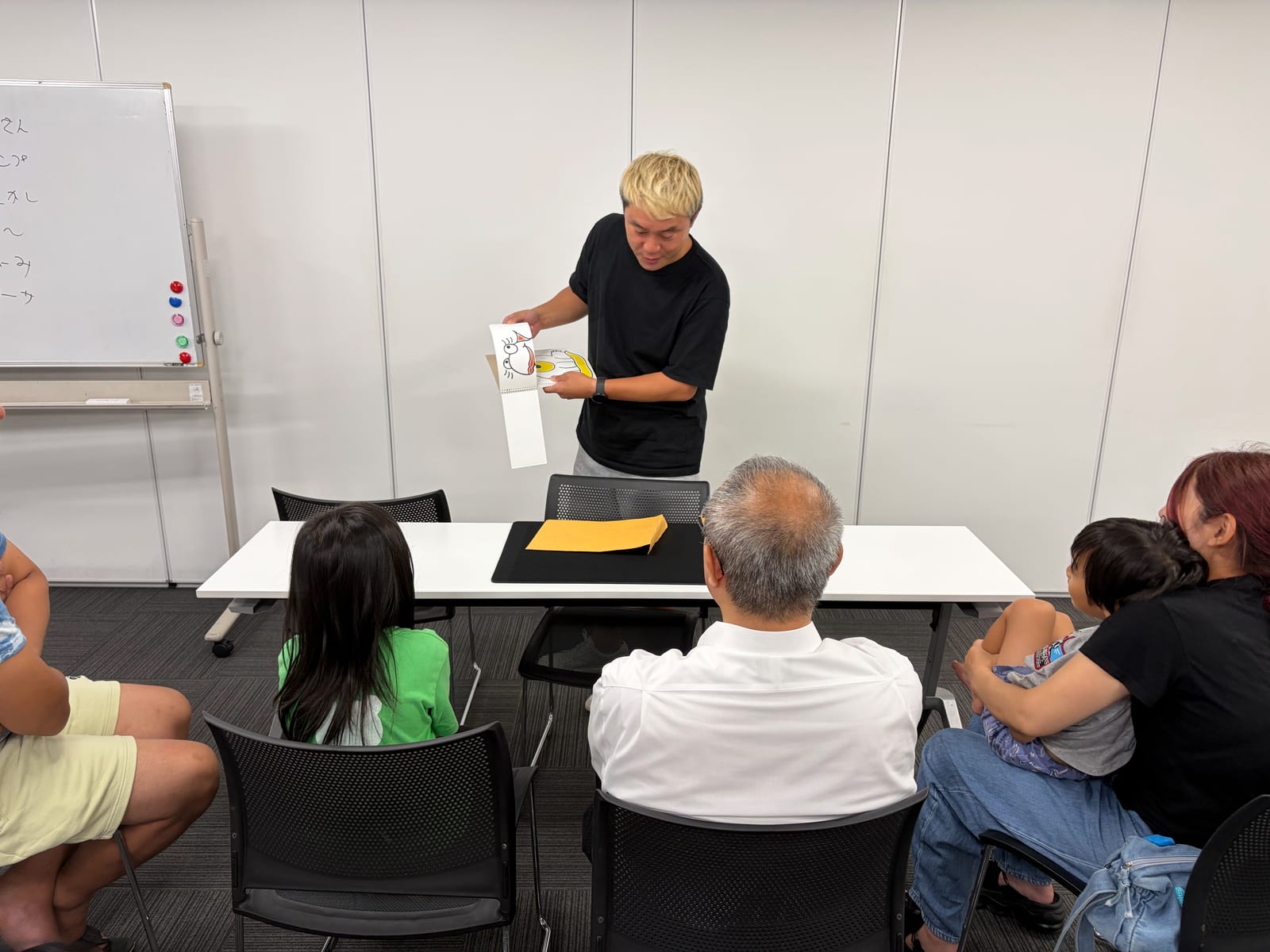 富山のマジックサークルを見学したご家族の夜|まじっぱ8月24日レポート 7 会場の輪の中で、金髪の若手メンバーがイラストマジックを見せる場面を後方から見学のご家族が見守る様子|富山マジックサークルまじっぱ