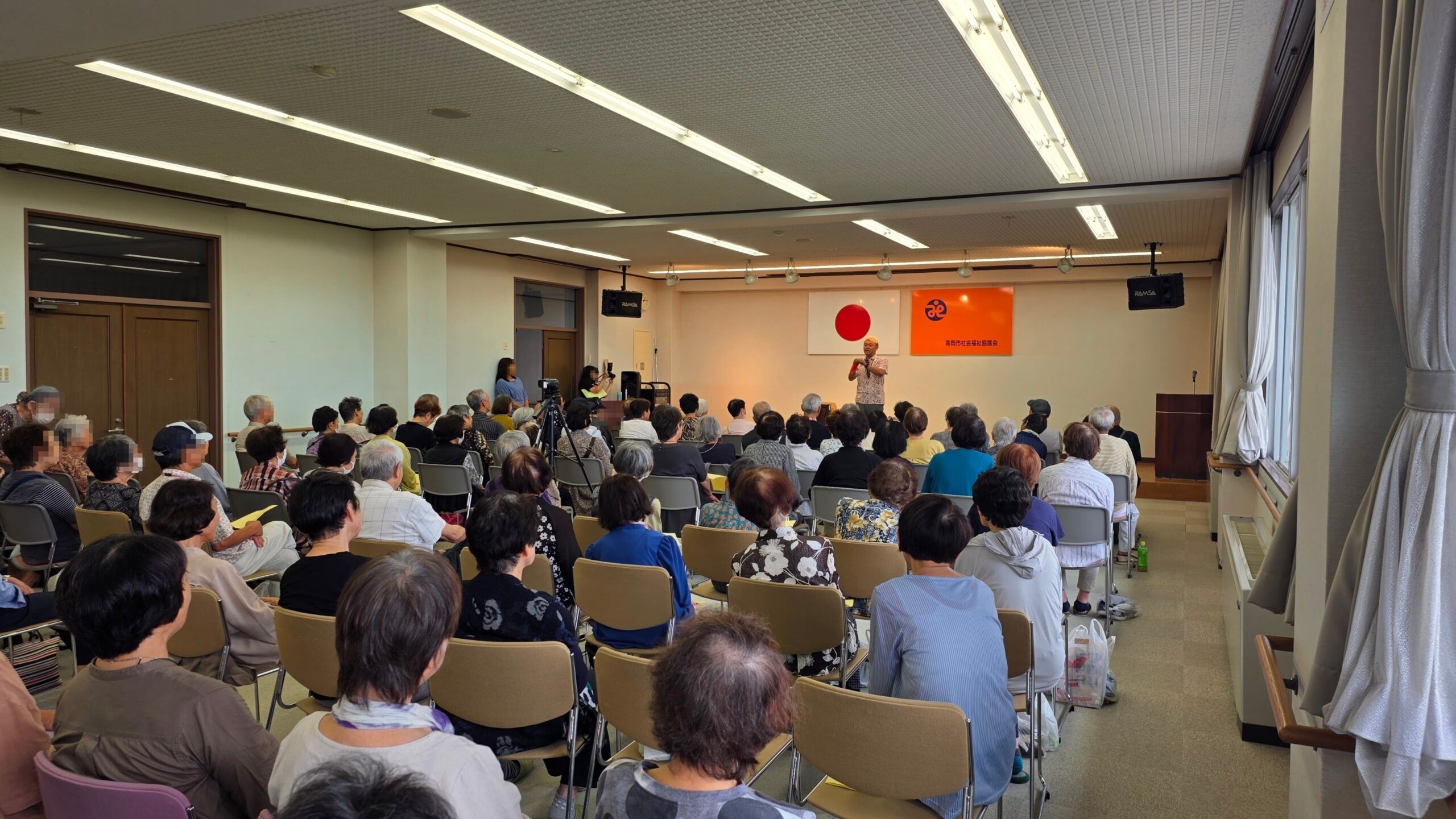 カードを手に高岡市の敬老会で客席へ語りかけるコンプレッサー
