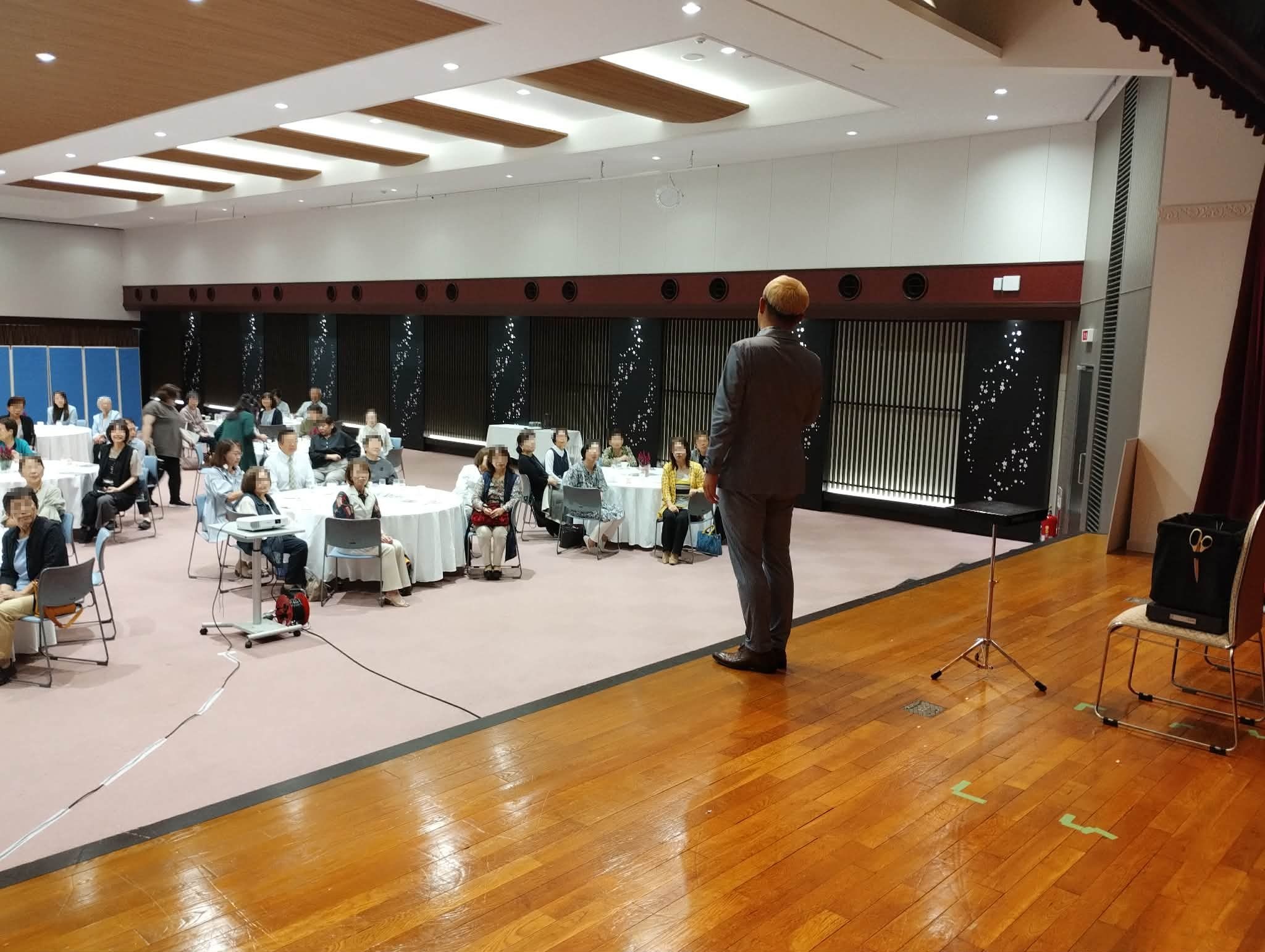高岡商工ビル2階大ホールで、円卓の女性客へ語りかけながらマジックショーを進める様子