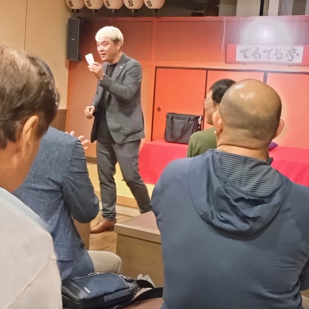 てるてる亭のミニシアターで、白いカードを手に客席へ話しかけるマジシャン・コンプレッサー
