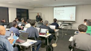 射水市の救急薬品市民交流プラザで開催、AIエージェント講座、満員御礼! 5 射水市で開いたAIエージェント講座で、スクリーンの横に立って話すコンプレッサーと、ノートパソコンを開いて聞く参加者の皆さん