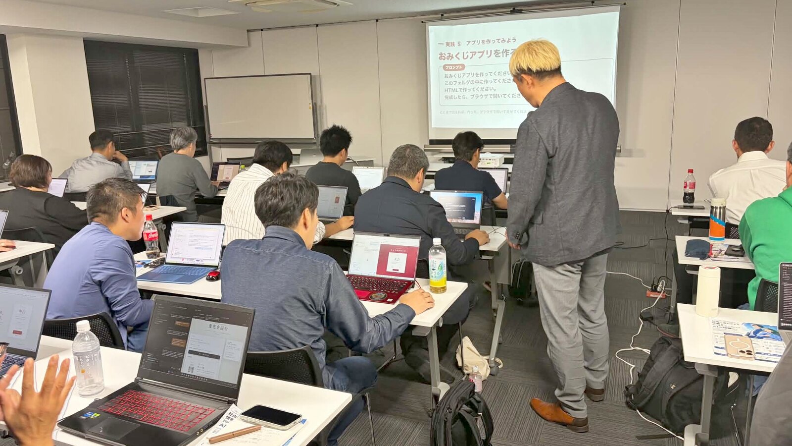 AIエージェント講座の実践の時間。スクリーンに「おみくじアプリを作る」と映り、参加者のパソコンにおみくじの画面が出ている。席のあいだを歩いて見て回るコンプレッサー