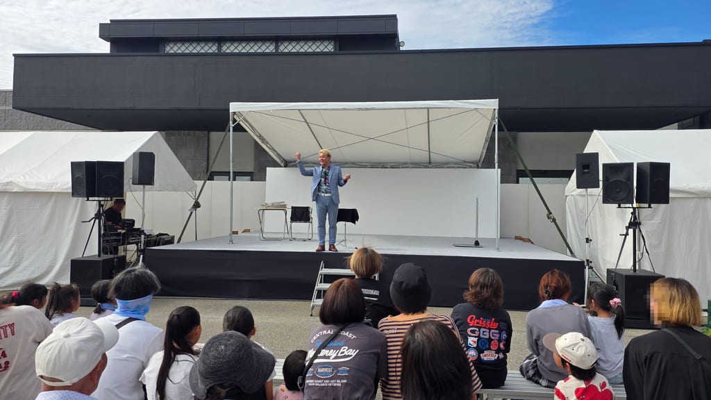朝日町のひすい野ホール前に組まれた屋外ステージで、両手のこぶしを上げるマジシャン・コンプレッサーと、ステージ前のベンチで見るお客さま