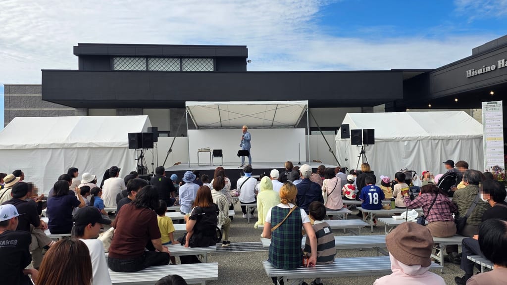 後ろから見た客席。ひすい野ホールの看板がある建物の前で、ベンチに座ったお客さまが屋外ステージのマジックショーを見ている