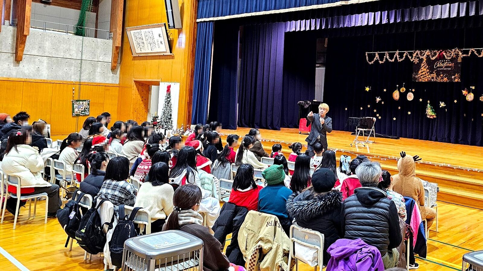 小学校の体育館で、クリスマスの飾りつけをしたステージの前に立ってマジックショーをするコンプレッサー（ダンス教室さんのクリスマスイベント）