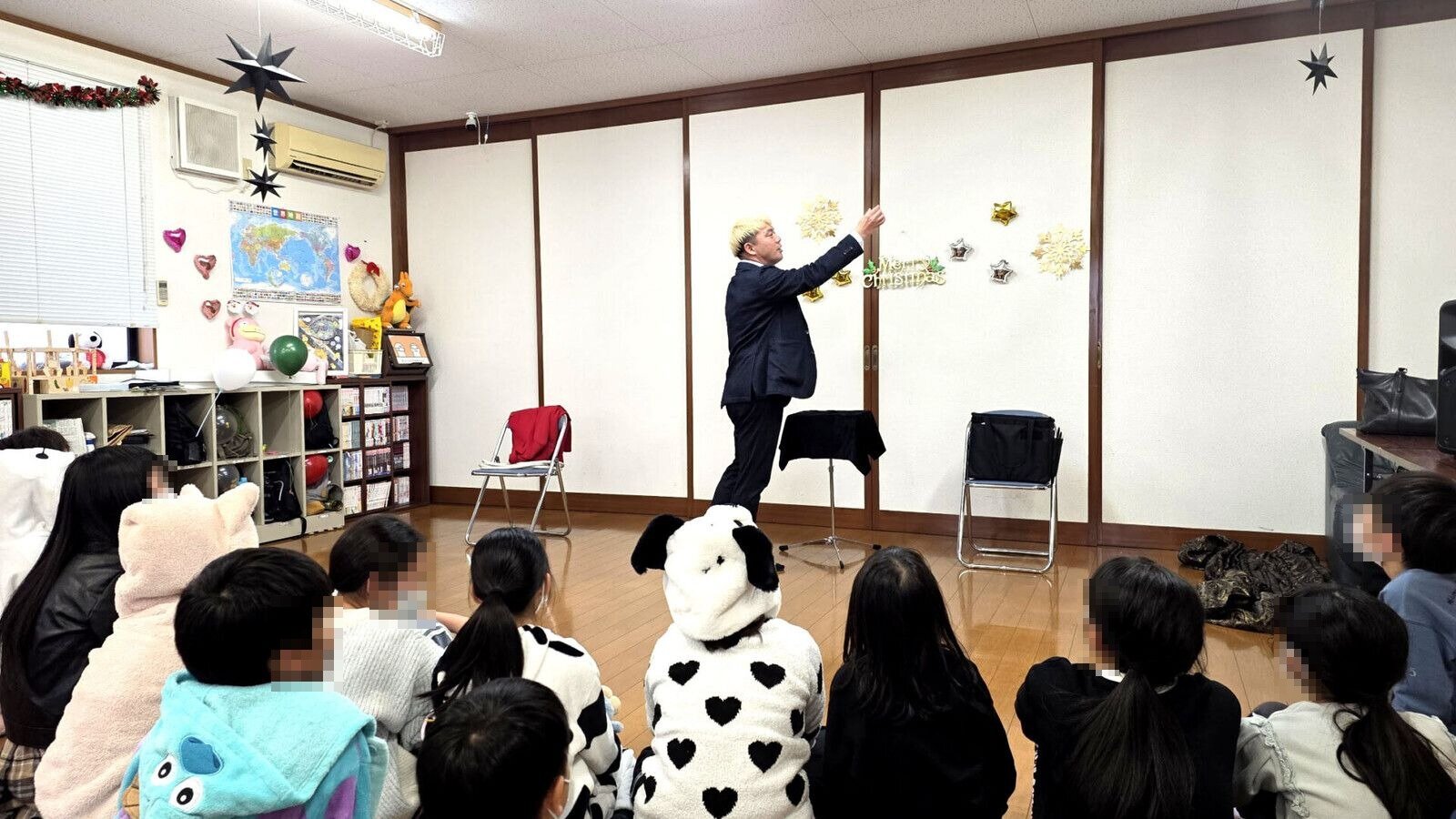 星やMerry Christmasの飾りの前でマジックを演じるコンプレッサーと、動物の着ぐるみ姿の子もいる客席（氷見市の学童のクリスマス会）