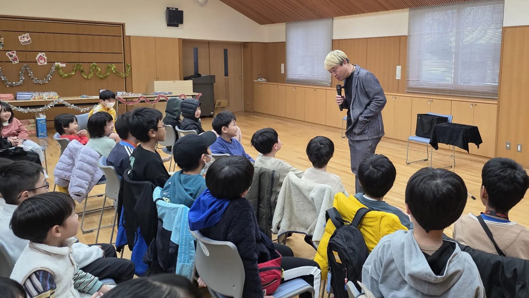 クリスマスの飾りつけをした児童クラブのクリスマス会で、子どもたちに語りかけながらマジックを進めるマジシャン コンプレッサー