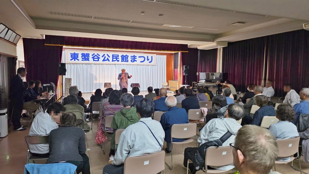 東蟹谷公民館の1階ホール。うしろの席から見たステージと、椅子に座ってマジックショーを見る地域の皆さん