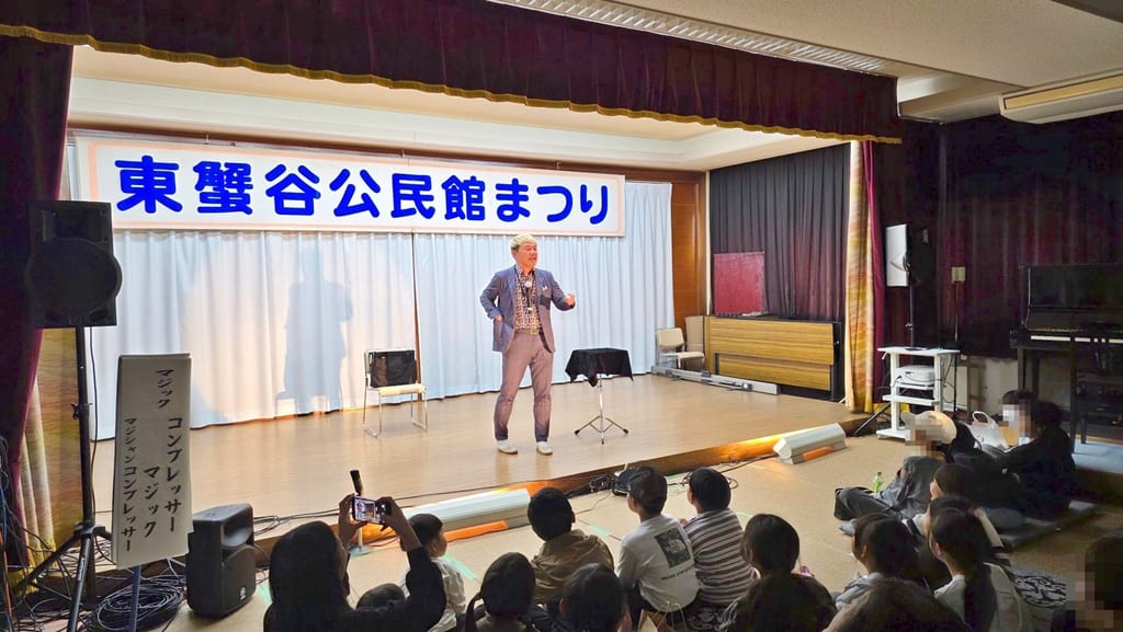 小矢部市の東蟹谷公民館まつりで、「東蟹谷公民館まつり」の横断幕の下のステージに立ち、前の床に座った子どもたちへ話しかけるコンプレッサー
