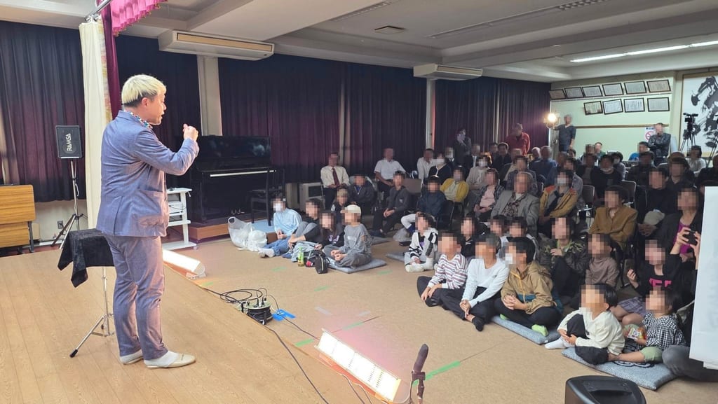 東蟹谷公民館の1階ホールで、ステージの端から客席へ語りかけるコンプレッサーと、前の床の座布団に座る子どもたち、うしろの椅子の席で見守る大人の皆さん