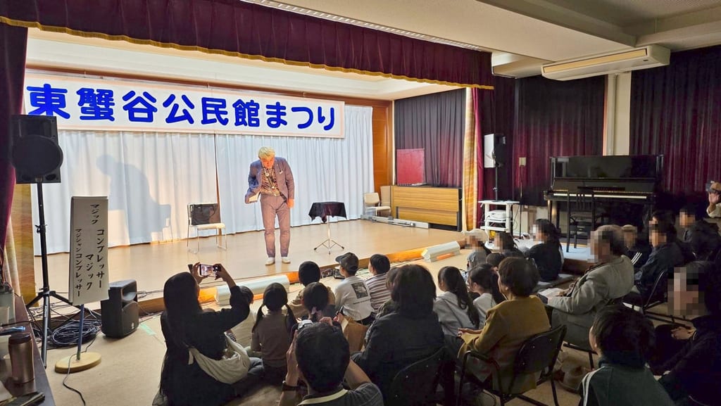 ステージの中央でおじぎをするコンプレッサーと、スマートフォンを向けてマジックショーを見る客席の皆さん