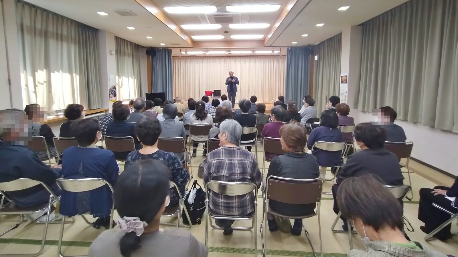 小矢部市の若林公民館で、畳の広間に椅子を並べた客席と、正面のステージに立つコンプレッサー