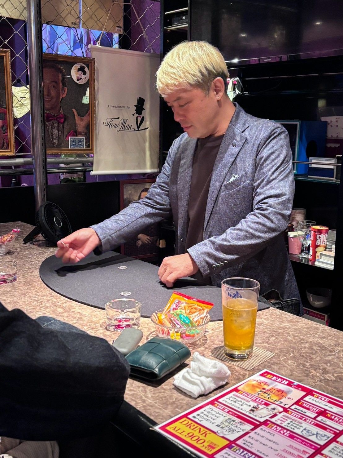 マジックバー ショーマンのカウンターで、銀貨を1枚つまんでお客さまに見せるコンプレッサー。うしろにShowManのタペストリーが見える