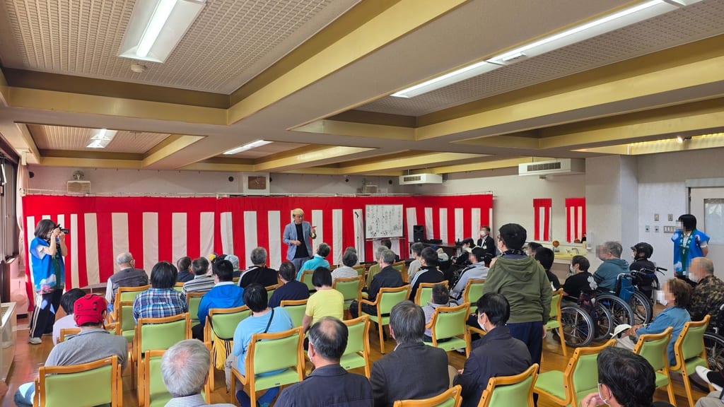志貴野苑祭の会場。紅白幕の前に立つコンプレッサーと、緑色のいすに座ってマジックを見る利用者の皆さん