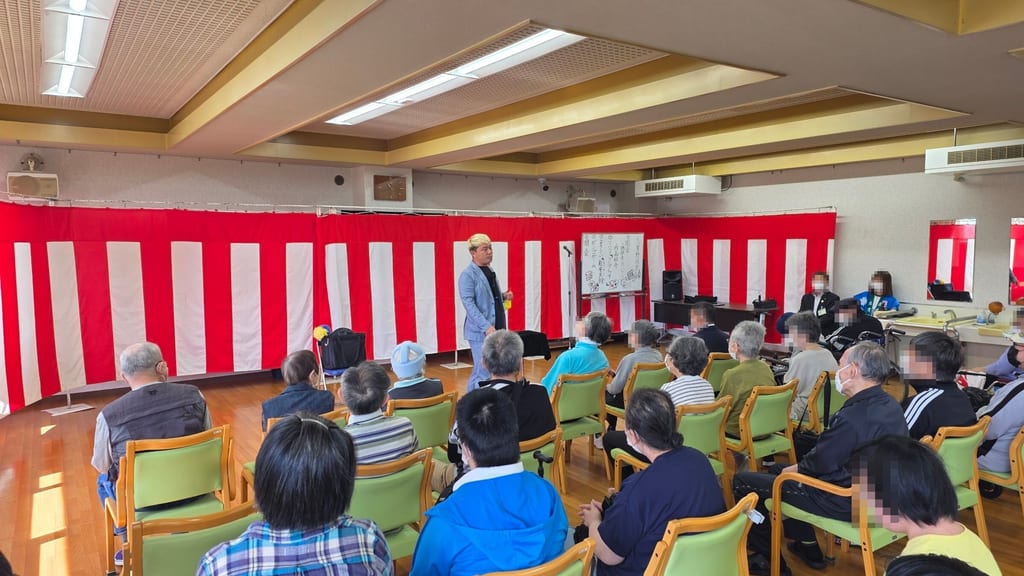 紅白幕の前に置かれた志貴野苑祭の手描きのプログラムと、その横で客席に話しかけるコンプレッサー