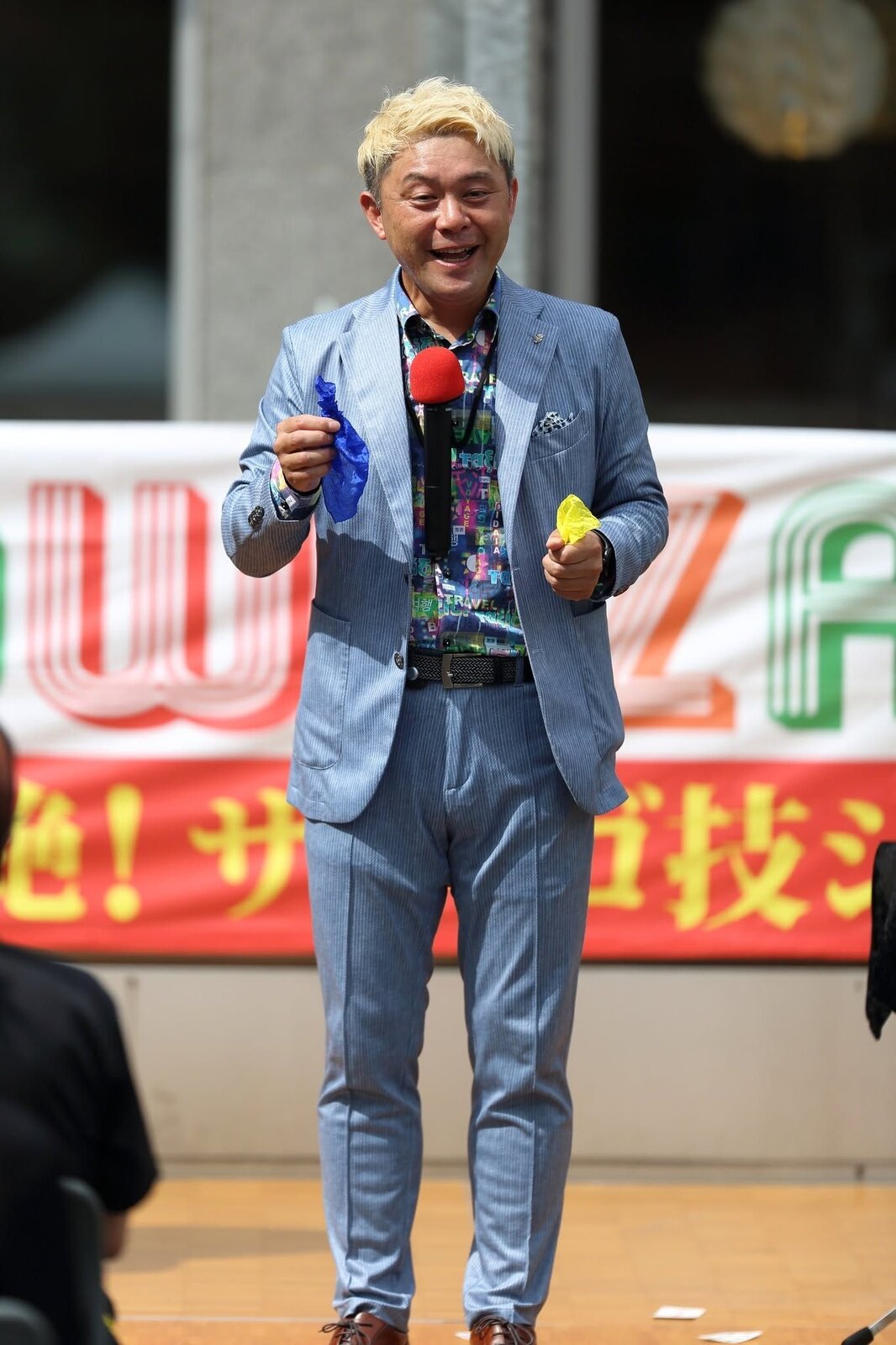 青と黄色のハンカチを両手に持ち、ハンカチの色を変えるマジックを演じるマジシャン・コンプレッサー（富山市民プラザ前庭）