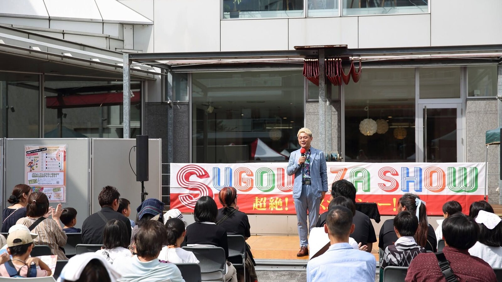 スゴ技ショーVOL81、富山市民プラザ前庭のステージでマイクを持って話すマジシャン・コンプレッサーと、客席の皆さん