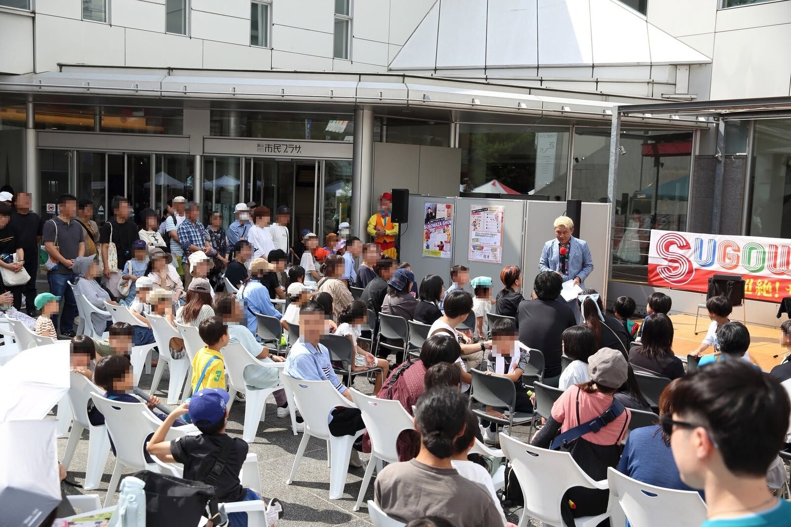 スゴ技ショーVOL81、富山市民プラザ前庭の客席の前で紙を手に話すマジシャン・コンプレッサーと、たくさんのお客さん