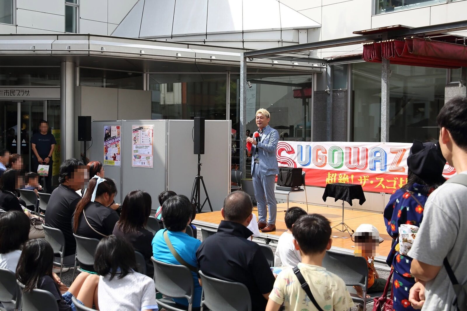 富山市民プラザ前庭のスゴ技ショーVOL81。SUGOWAZAの横断幕の前でマジックショーをするマジシャン・コンプレッサーと、見守る親子連れ