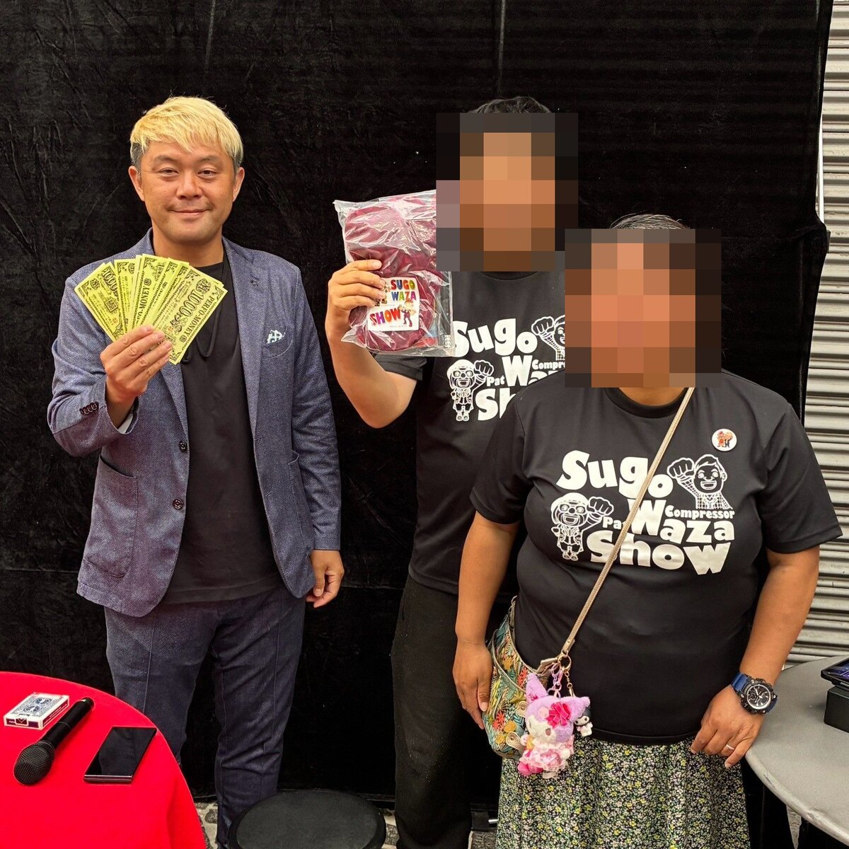 スゴ技ストリートで、パフォマネーの束を広げるコンプレッサーと、景品を掲げる常連のお客さんおふたり。どちらもスゴ技ショーのTシャツ姿