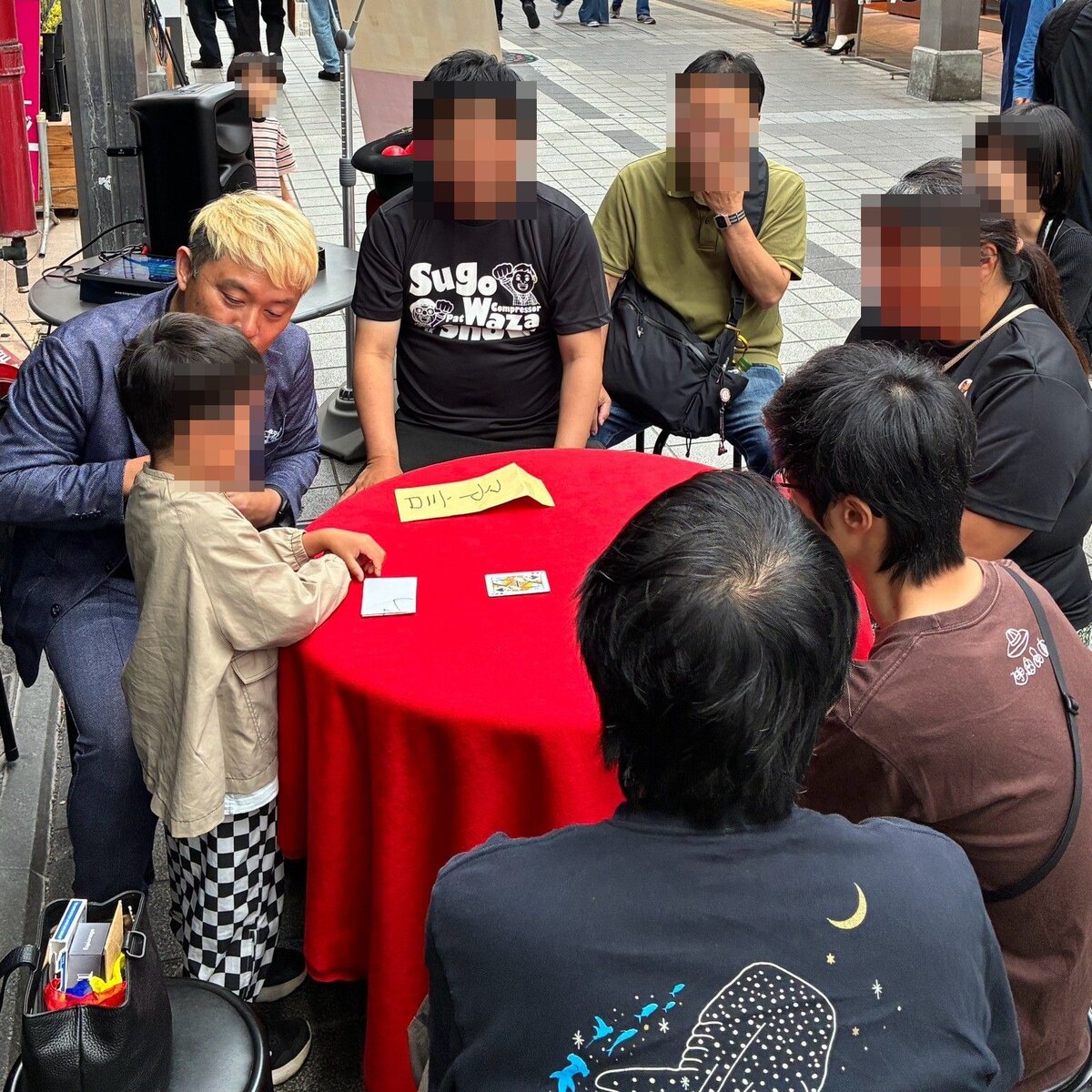 スゴ技ストリートで、男の子のとなりに座ってトランプの予言のマジックをするコンプレッサー。赤いテーブルの上に黄色い予言の紙が置いてある