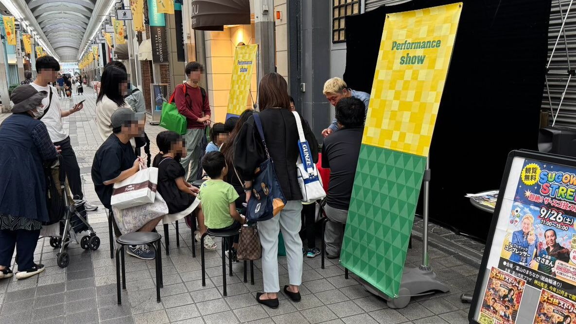 富山・総曲輪通りのスゴ技ストリート。9月26日の看板の横で、テーブルを囲むお客さんにテーブルマジックを見せるマジシャン・コンプレッサー