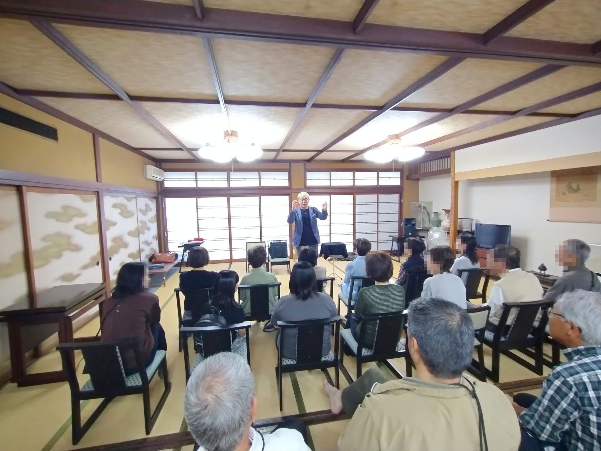 高岡市福岡町の料理旅館の広間に椅子を並べた客席と、両手を上げて話すコンプレッサー